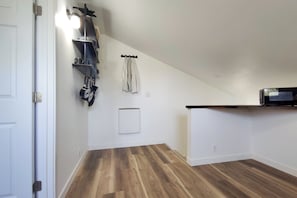 A room with a wooden floor, a white door, a black shelf with kitchen items, a white radiator, and a microwave on a counter.