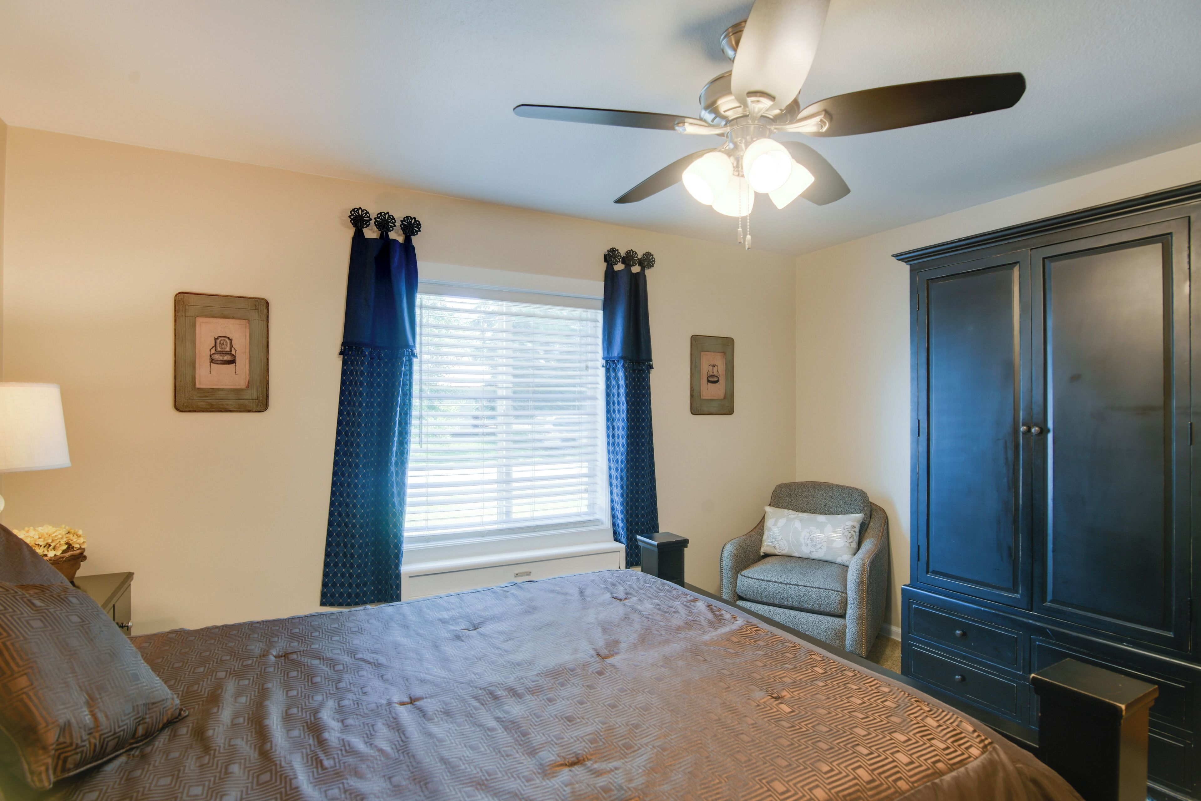 Une chambre à coucher comprenant un lit, un ventilateur de plafond, une fenêtre avec des rideaux, une chaise et une armoire.