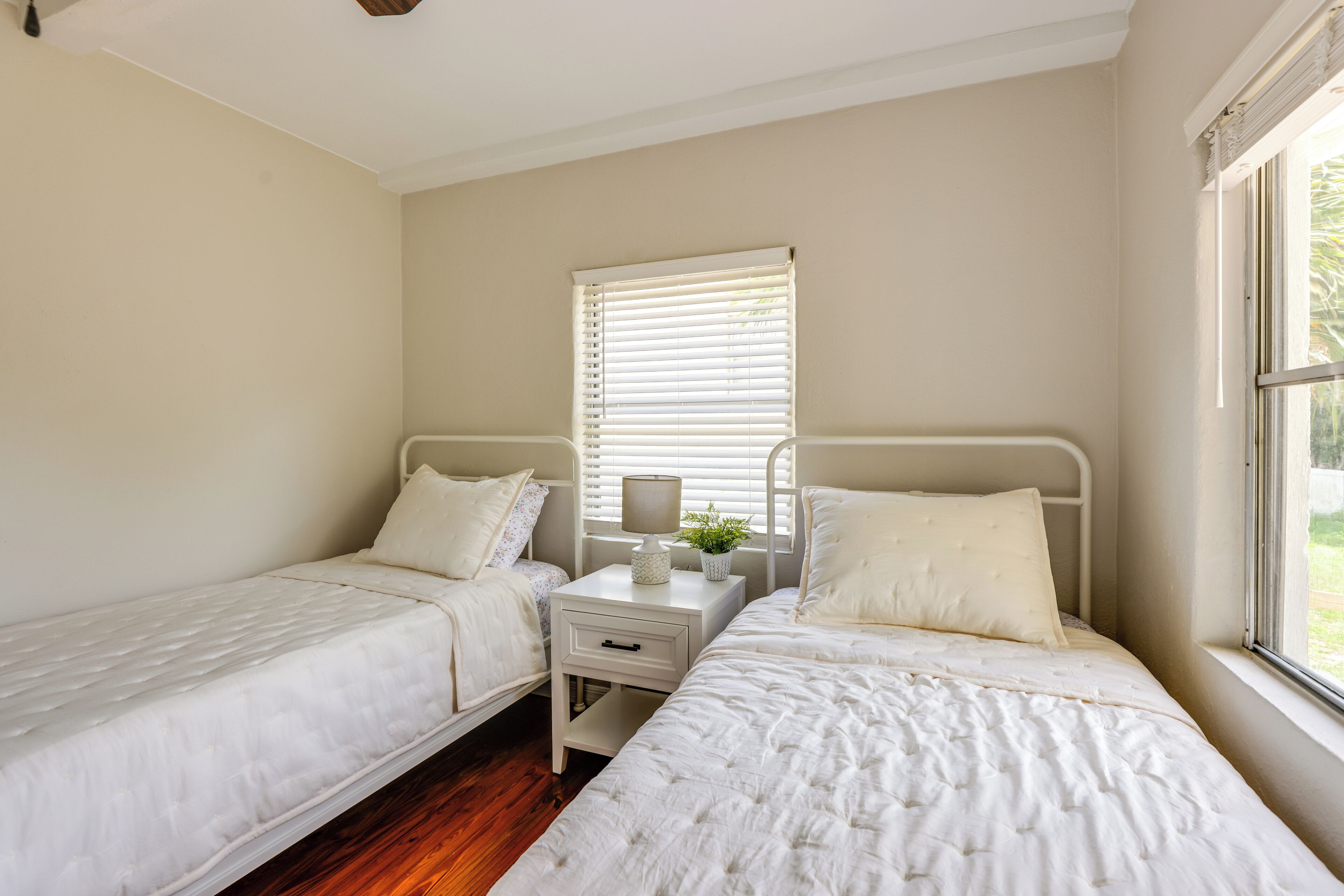 A bedroom with two single beds, a bedside table with a lamp, a window with blinds, and a small plant on the table.