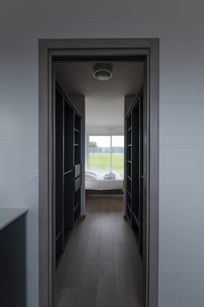 Ein schmaler Flur, der zu einem Schlafzimmer mit großem Fenster, Holzboden und integrierten schwarzen Regalelementen führt.