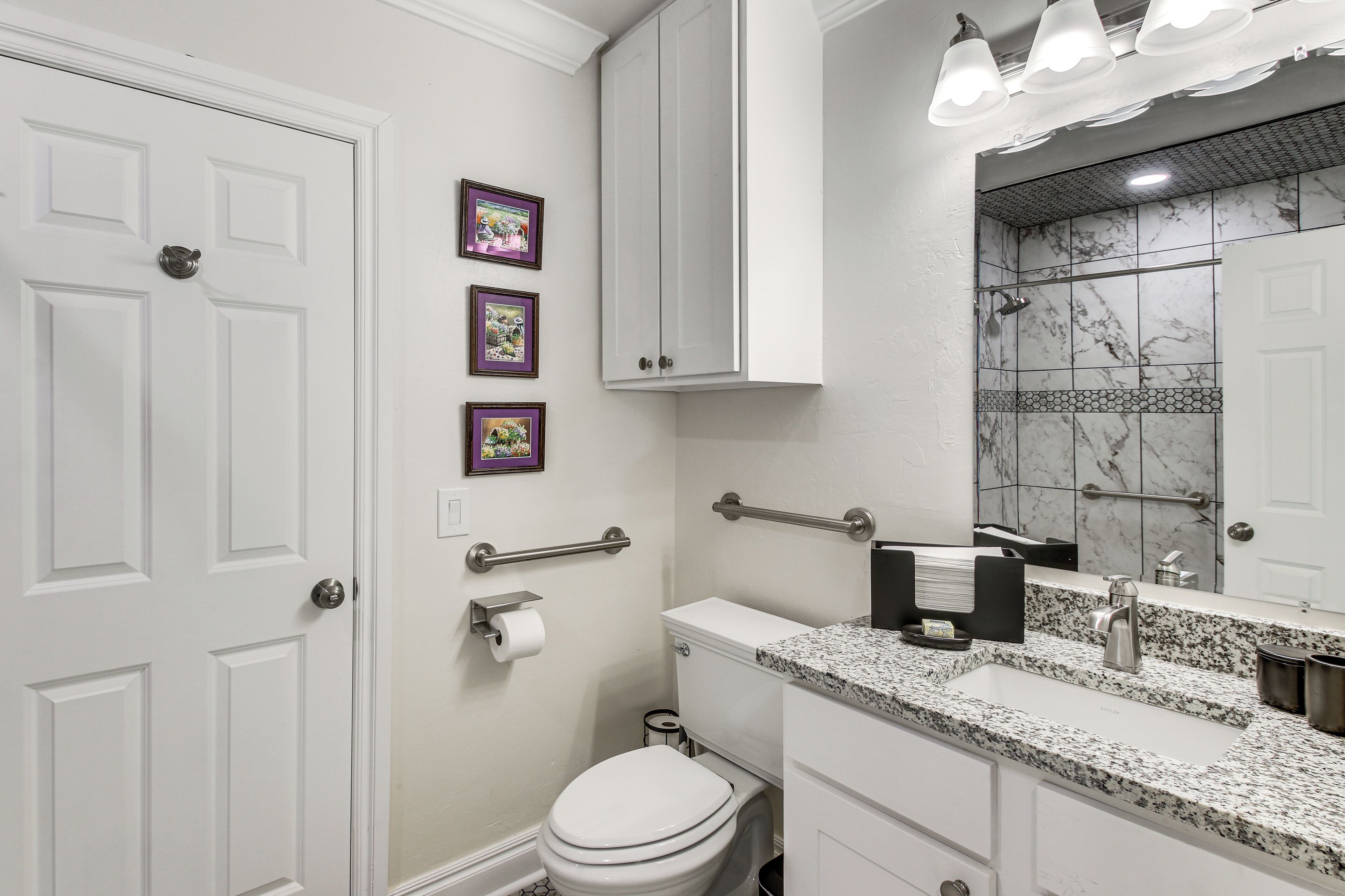 Une salle de bain avec des toilettes blanches, un lavabo doté d’un plan de travail en granit, un miroir avec un pare-feu en marbre et une porte blanche.