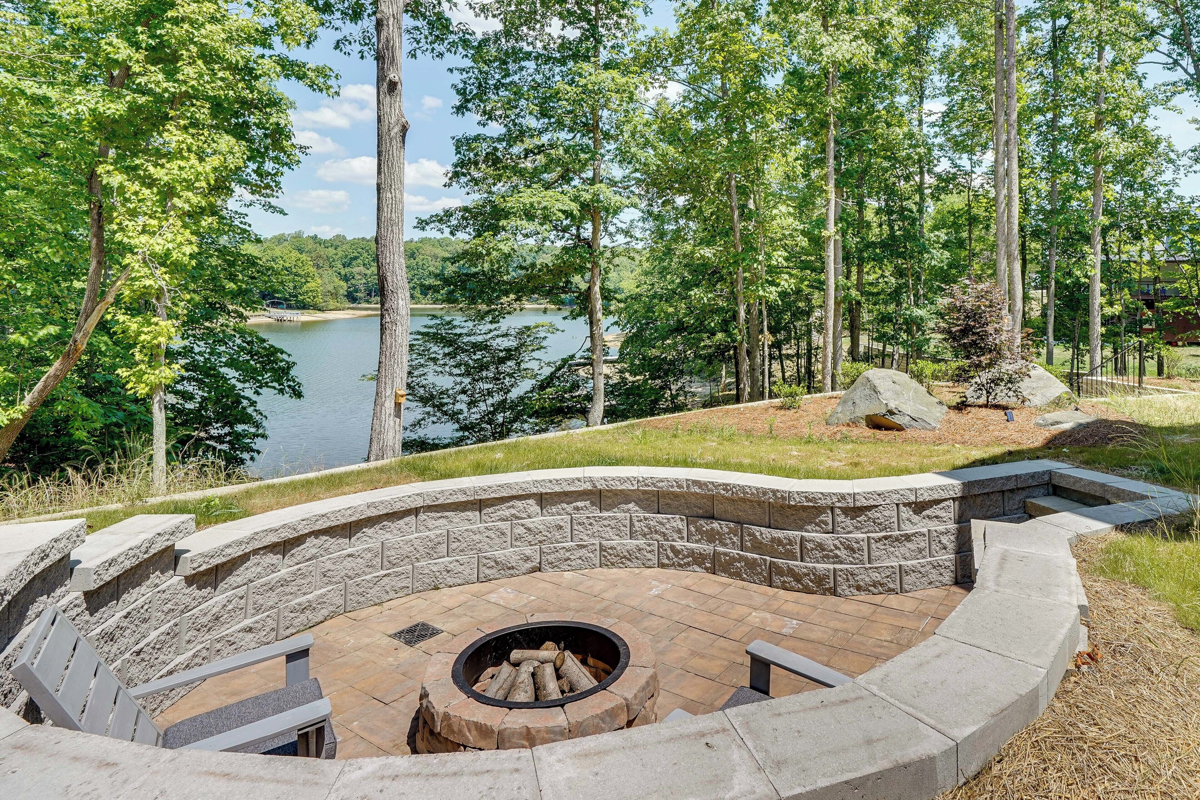 A stone fire pit surrounded by a curved stone bench, overlooking a lake with trees in the background.