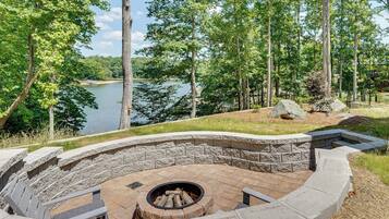 A stone fire pit surrounded by a curved stone bench, overlooking a lake with trees in the background.