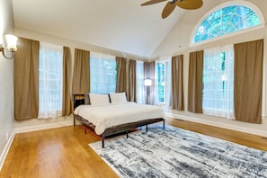 Un dormitorio con una cama, ventilador de techo, ventana grande con cortinas y piso de madera.