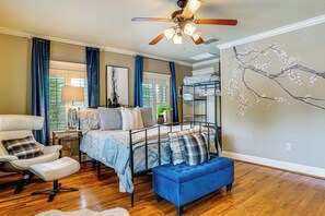 A bedroom with a bed, a chair, a blue bench, a ceiling fan, and a wall with a tree painting.