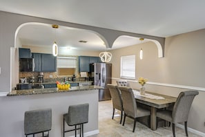 A modern kitchen with granite countertops, a stainless steel refrigerator, and a dining area with a wooden table and gray upholstered chairs. - Golf, Beaches & Sunshine! Pet-friendly Naples Home (Naples)