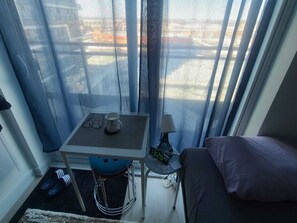 Ein kleines Zimmer mit Tisch, Stuhl, Bett und einem Fenster mit Vorhängen.