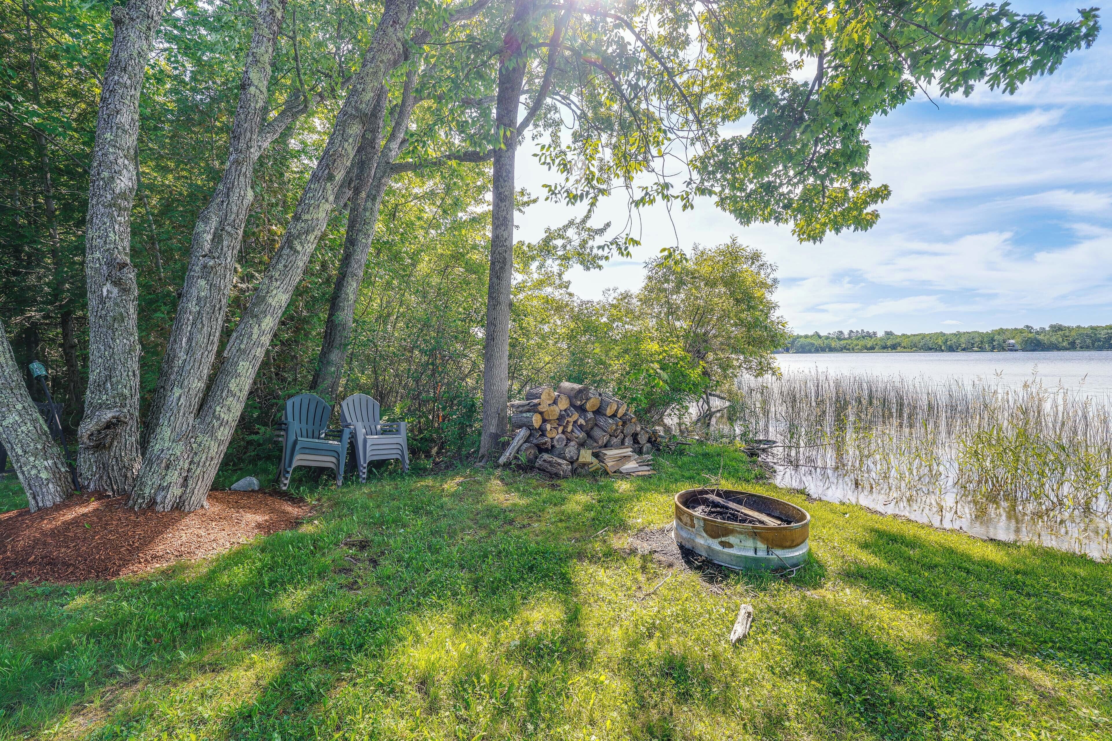 A lakeside setting with trees, a pile of logs, two Adirondack chairs, and a canoe.