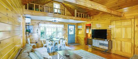 Interior de una cabaña de madera con comedor, sofá, televisor y habitación de lecho superior.