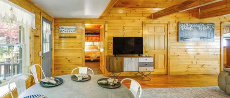 A wooden cabin interior with a dining table set for four, a television, and a painting on the wall.