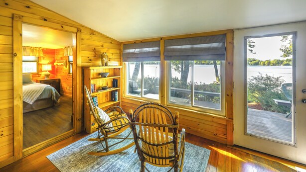 A wooden cabin interior with a bed, rocking chairs, a bookshelf, and large windows offering a view of the outdoors.