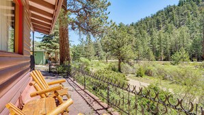 A wooden deck with outdoor seating, a metal fence, and a forested hillside in the background. - On-site Fishing! Riverfront Gem w/ Patios in Drake (Drake)