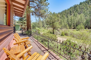 A wooden deck with outdoor seating, a metal fence, and a forested hillside in the background. - On-site Fishing! Riverfront Gem w/ Patios in Drake (Drake)