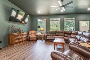 Ein Wohnzimmer mit Ecksofa, Holzschrank, Deckenventilator und großen Fenstern.