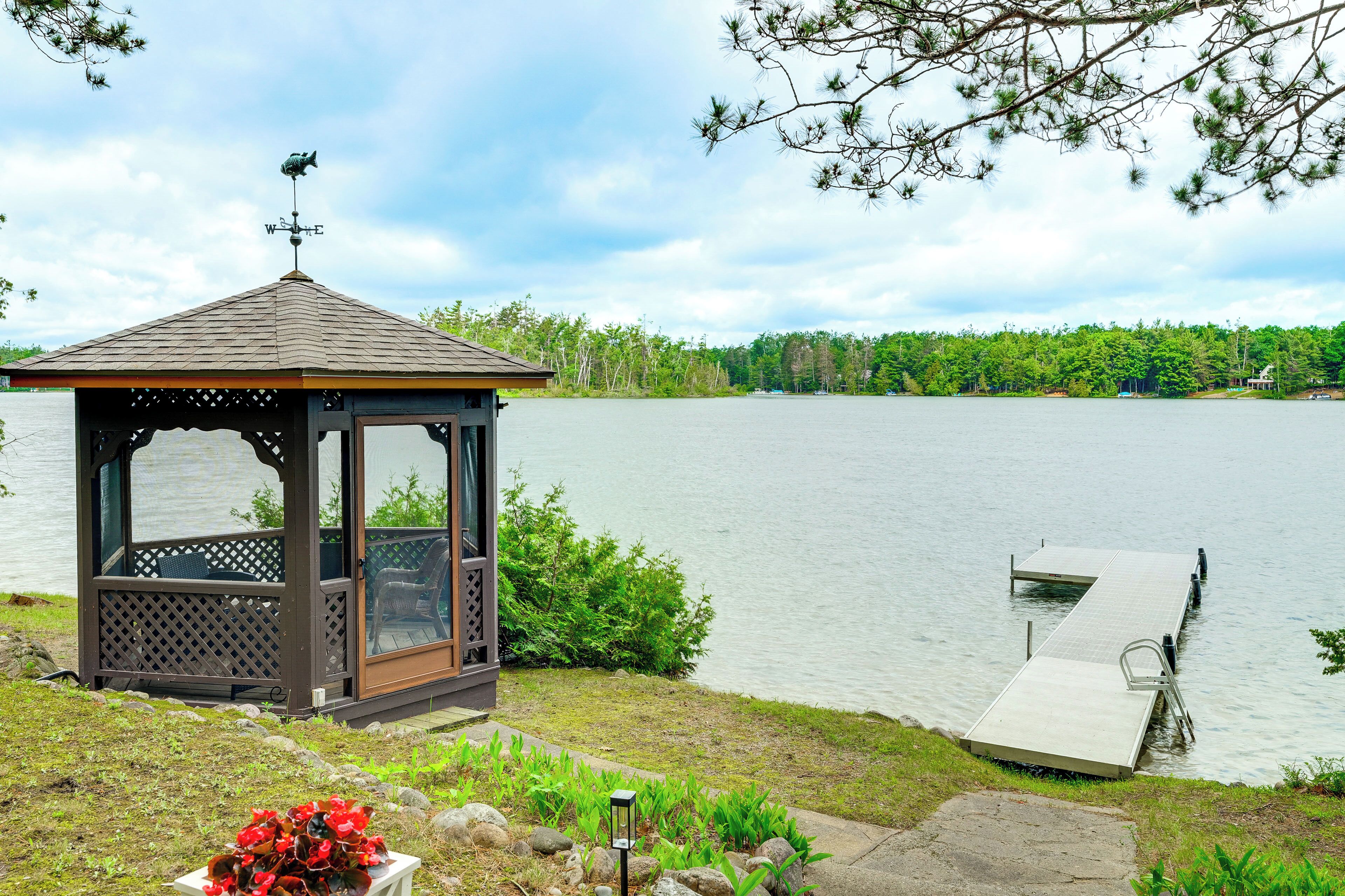 A gazebo with a roof and lattice panels, a dock extending into a lake, and a view of a forested shoreline.