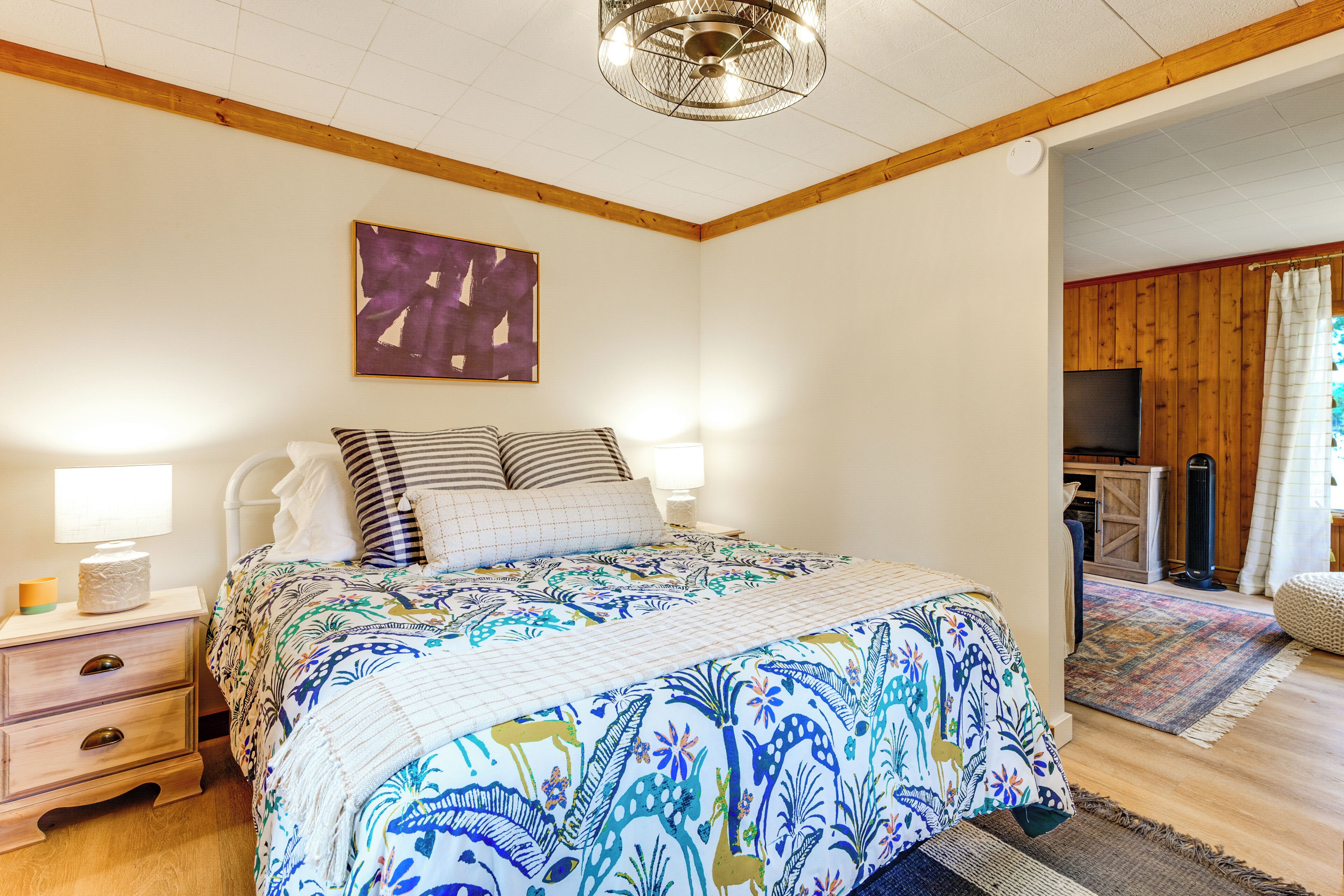A bedroom with a bed, bedside lamps, a nightstand, a wooden ceiling, a wall-mounted TV, and a door leading to another room.