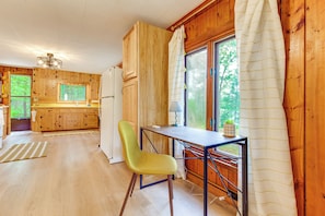 A wooden cabin interior with a dining table, a chair, a window with curtains, and a kitchen area. - Waterfront Tee Lake Cabin: Dock, Fire Pit + More! (Lewiston)
