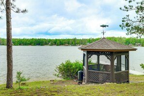 Ein Pavillon am See mit einer Metall-Fischskulptur darauf, umgeben von Bäumen und einem bewölkten Himmel.