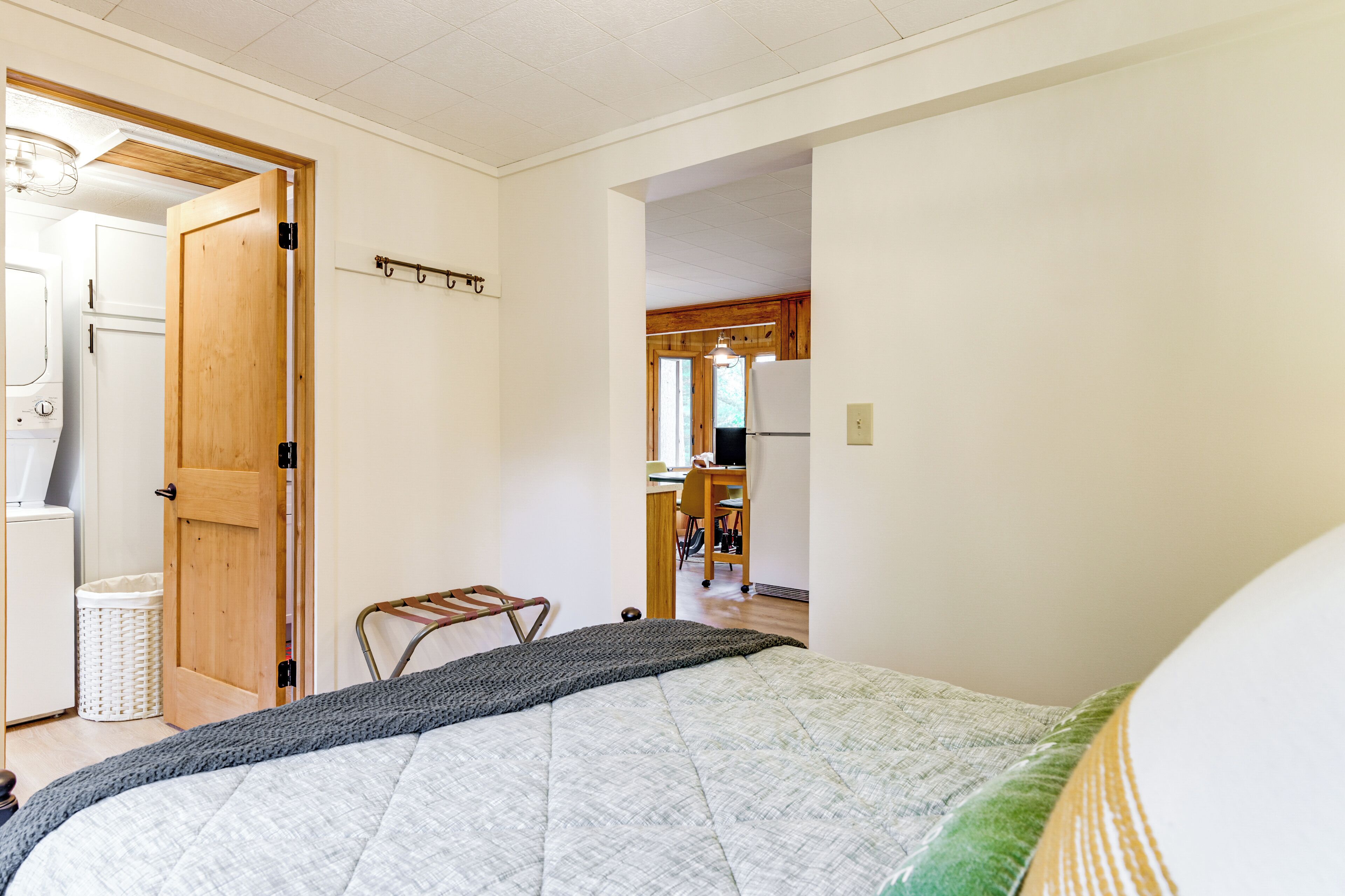 A bedroom with a bed, a wooden door, a laundry basket, and a view into the kitchen.