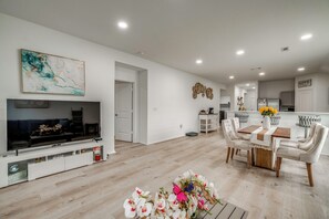 A modern living room with a flat-screen TV, a dining area with a wooden table and white chairs, and a kitchen in the background.