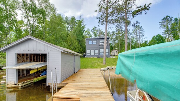 Un muelle de madera que lleva a un cobertizo pequeño con un canoa y un kayak, un edificio moderno de dos pisos al fondo, y una lancha cubierta.