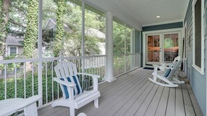 Eine überdachte Veranda mit weißen Geländern, zwei Adirondack-Stühlen und Blick auf ein Haus und Bäume.