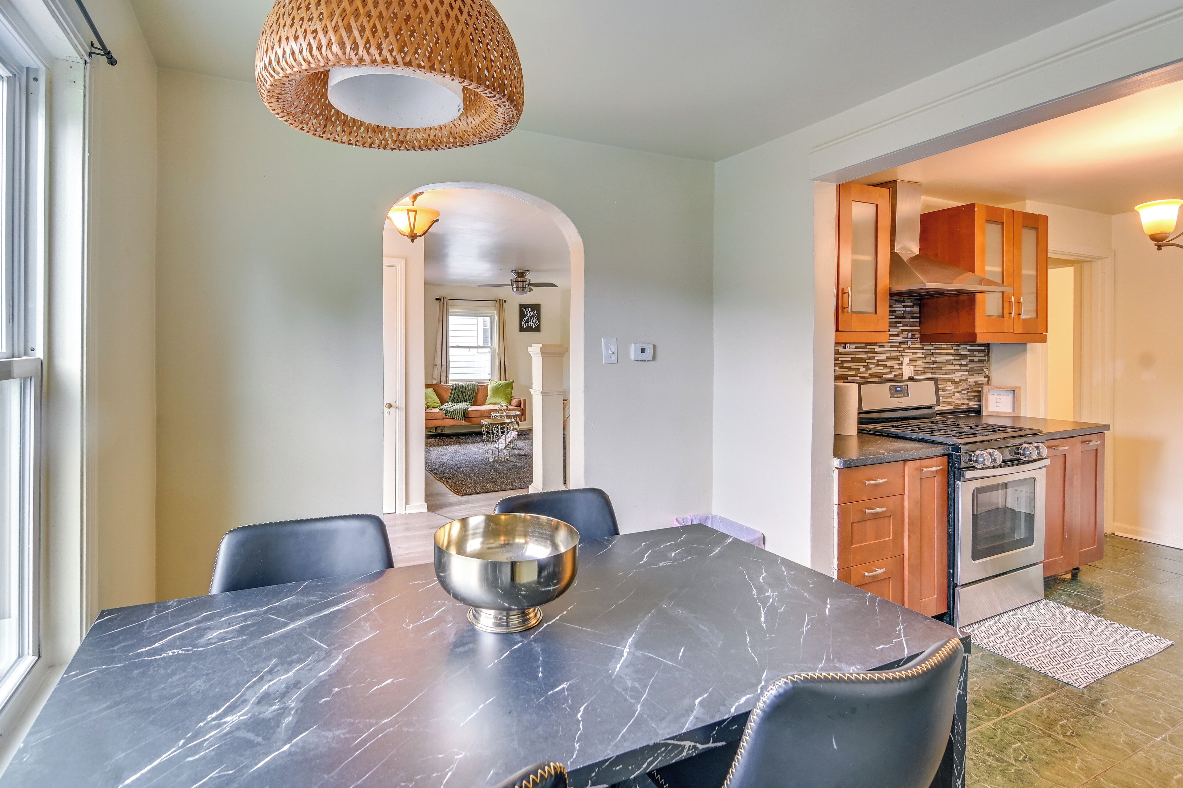 A dining area with a marble table, black chairs, and a silver bowl. There is a kitchen with wooden cabinets and a stove.