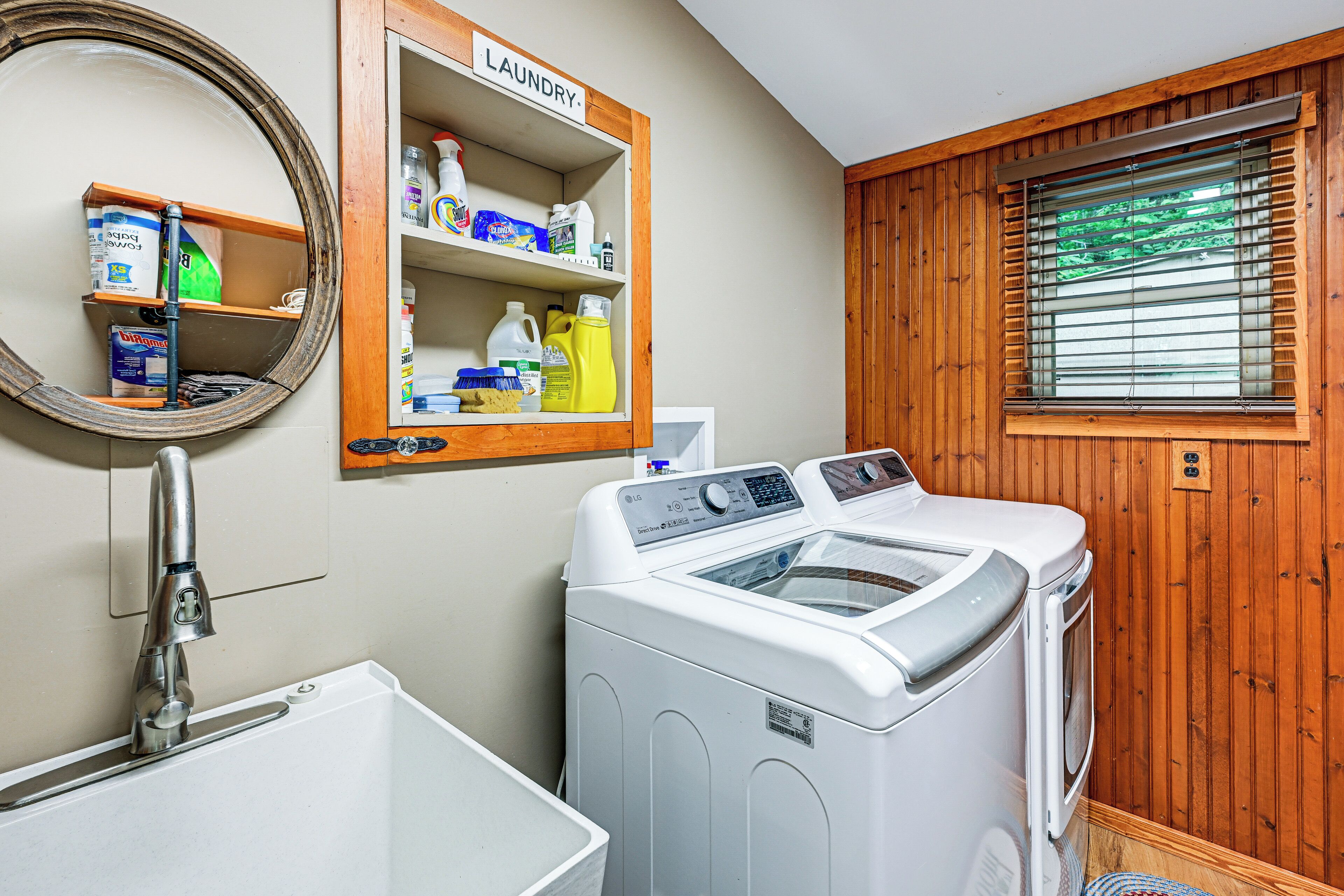 A laundry area with a washing machine, a shelf with cleaning supplies, a mirror, and a window with blinds.