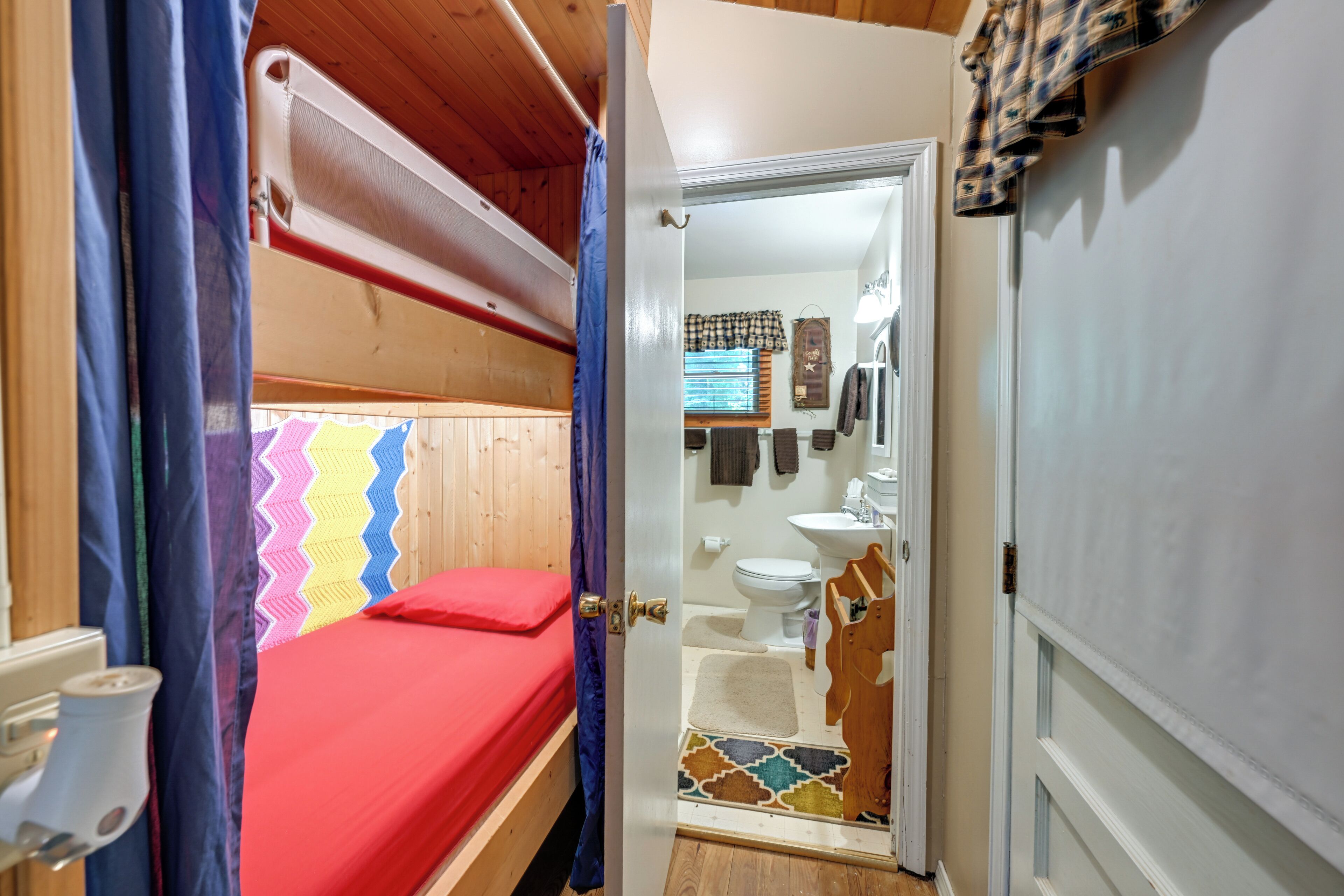 A bunk bed with a red mattress, a bathroom with a toilet, sink, and towel rack, and a door leading to another room.