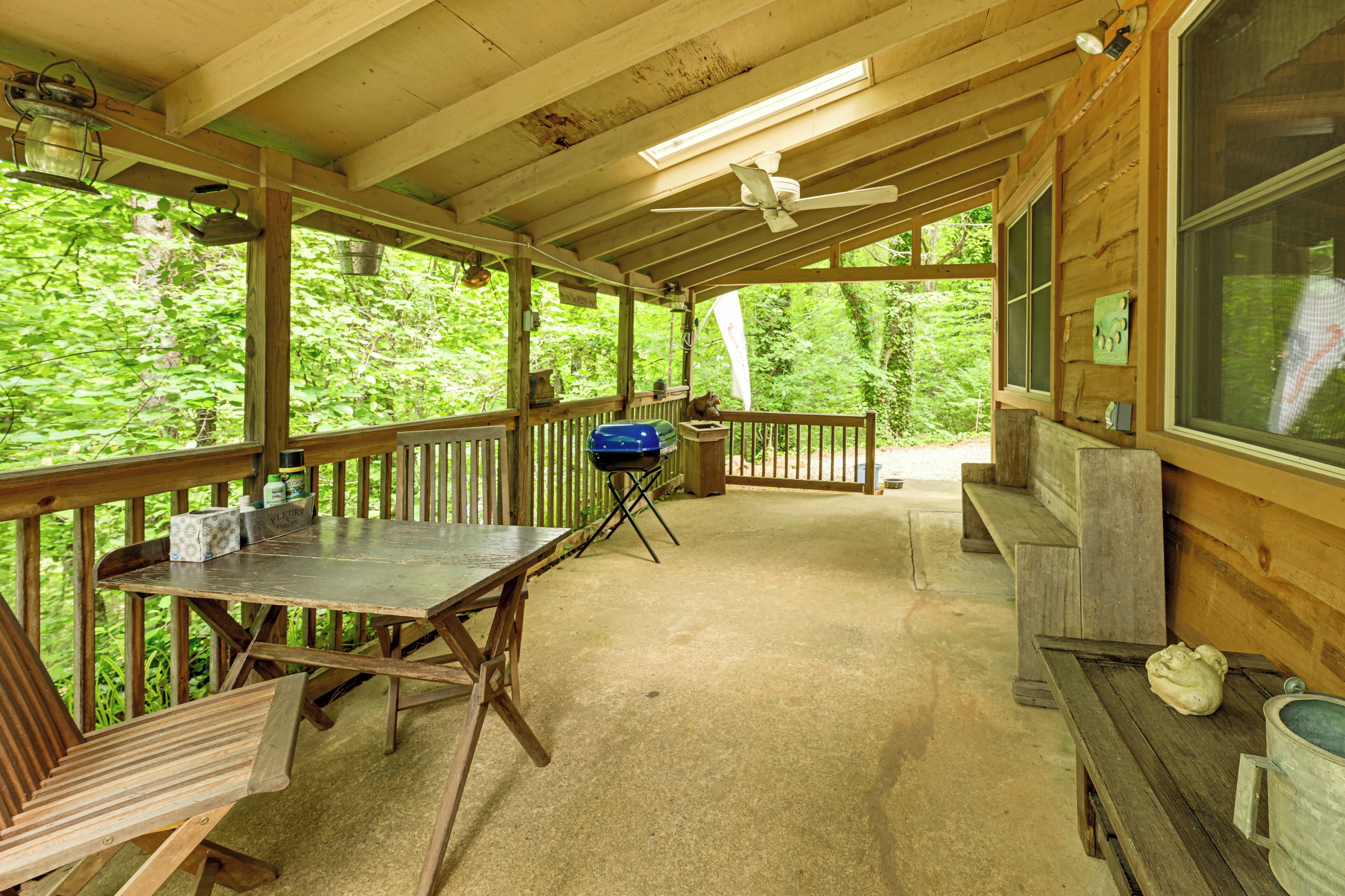 A covered outdoor area with a wooden table, benches, a grill, and a view of the surrounding forest.
