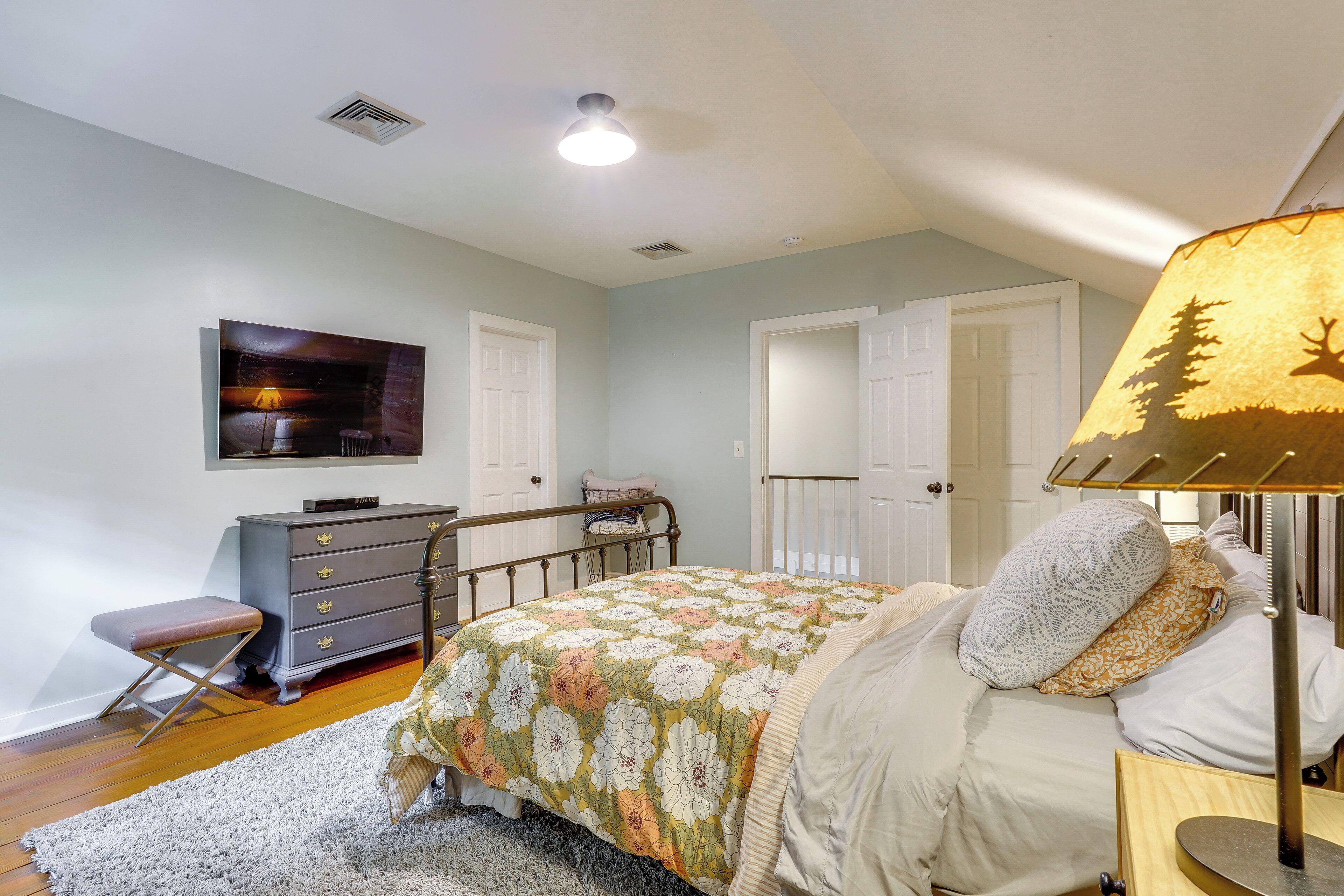 A bedroom with a bed, a dresser, a bench, a lamp, and a television on the wall.