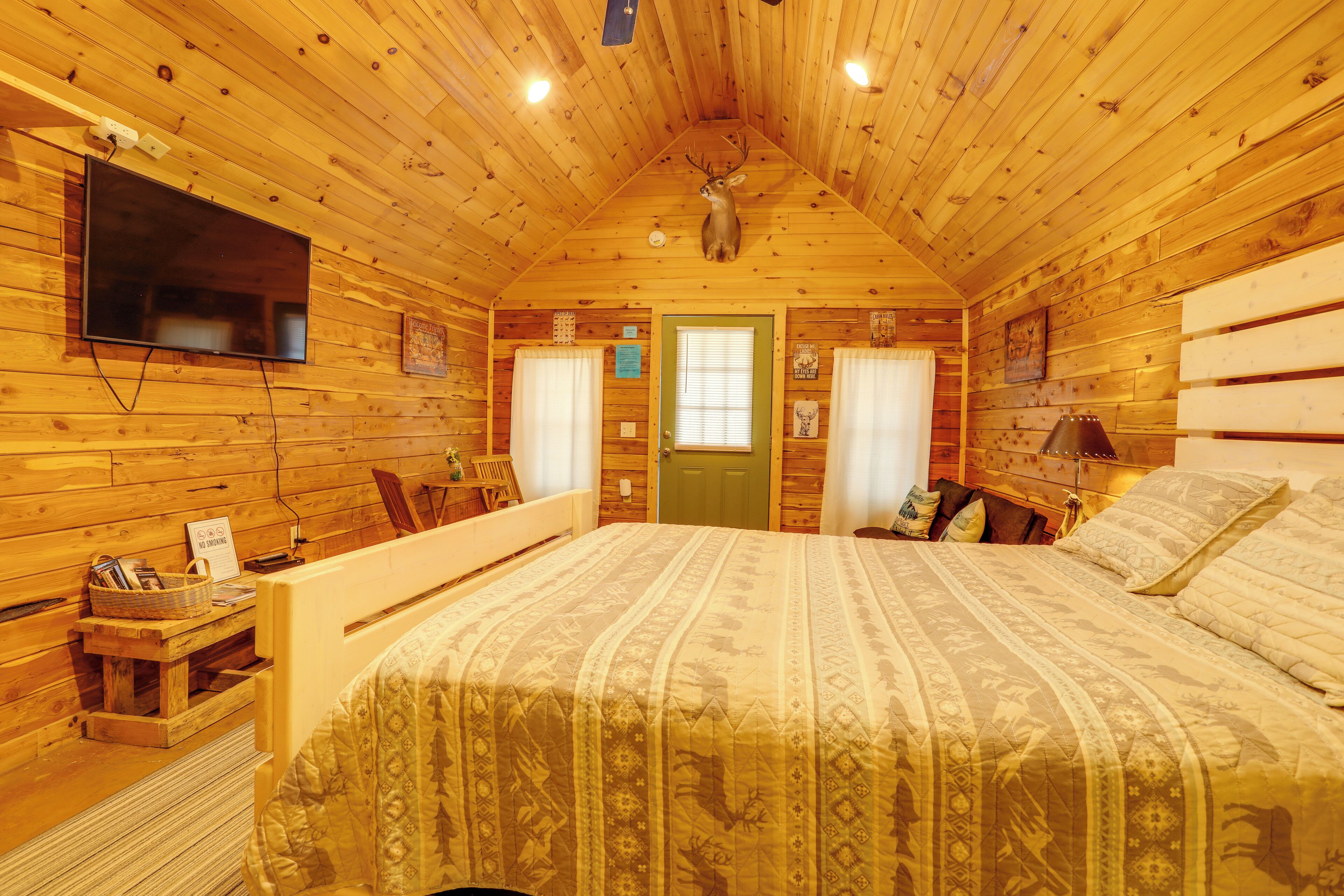 A wooden cabin bedroom with a bed, a mounted television, a deer head on the wall, and a window with curtains.