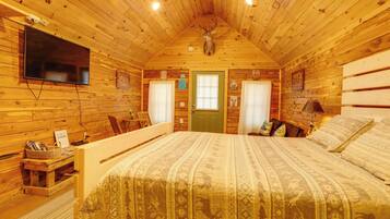 A wooden cabin bedroom with a bed, a mounted television, a deer head on the wall, and a window with curtains.