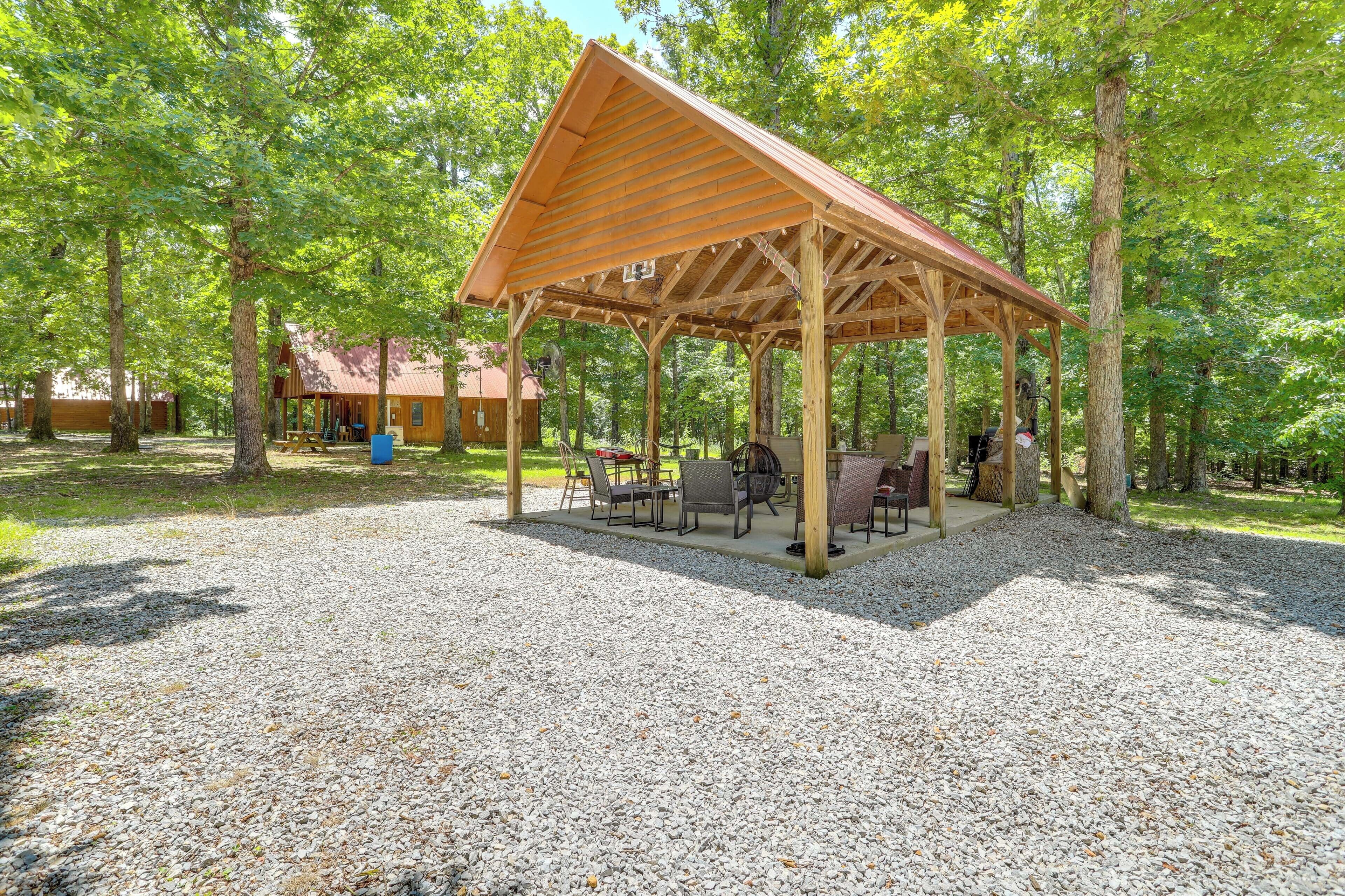 A covered outdoor seating area with tables and chairs, surrounded by trees.