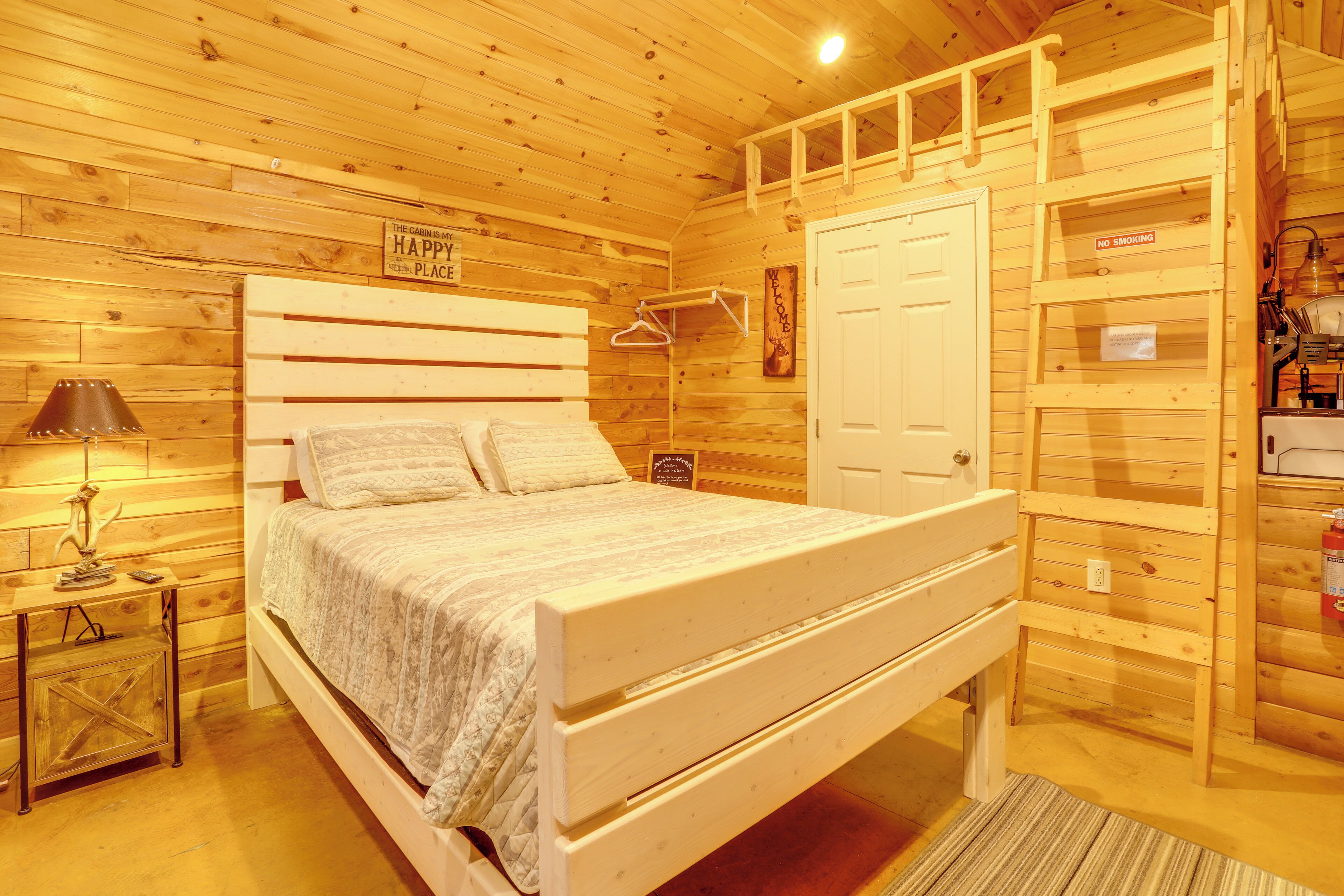 A wooden cabin-style room with a bed, a nightstand, a lamp, a coat rack, and a door with a 'NO SMOKING' sign.