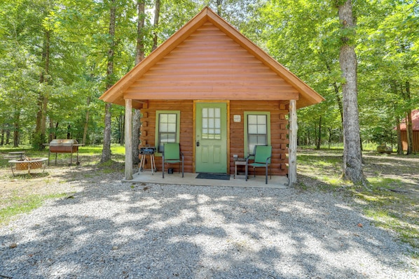 Une petite cabane en rondins avec une porte verte, deux fenêtres et une véranda avec des chaises.
