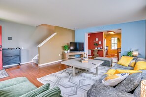 A living room with a sofa, coffee table, and TV. There is a staircase and a kitchen area visible. - Pet-friendly Home w/ Private Yard Near Pittsburgh (Clairton)