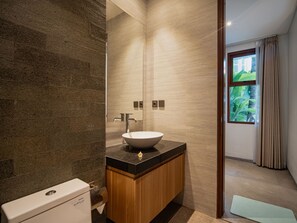 A modern bathroom with a stone wall, a wooden vanity, a white sink, and a toilet. - Villa RL Sanur (Sanur)
