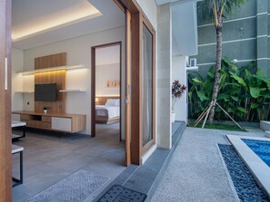 A modern hotel room with a bed, bedside table, TV, and a view of a swimming pool and tropical plants. - Villa RL Sanur (Sanur)