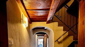 Un pasillo estrecho con una escalera de madera a la derecha, una pared de piedra a la izquierda y una vista hacia otra habitación al final.