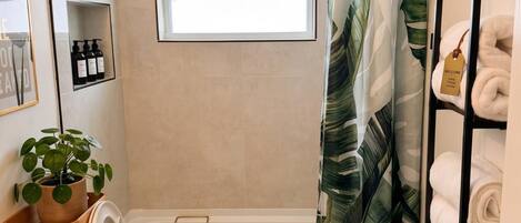 Baño con inodoro, ducha con cortina, ventana, repisa con artículos de aseo y toallero.