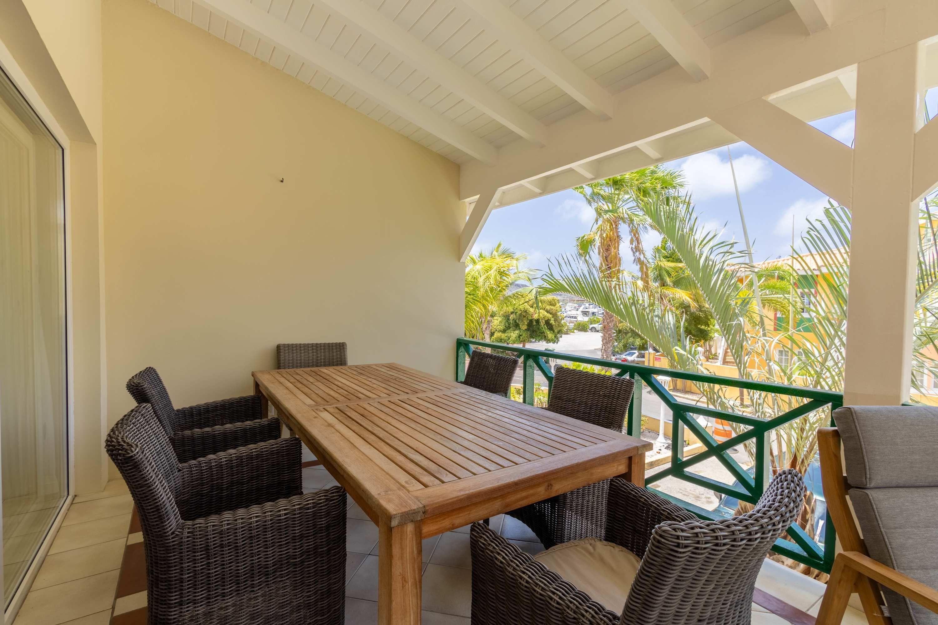 Un patio cubierto con una mesa de madera y sillas de mimbre, una barandilla metálica verde y vistas a palmeras y edificios.