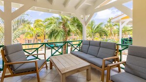 A covered outdoor seating area with a wooden table and grey cushioned chairs, overlooking palm trees and buildings. - Spanish Water Marina Superior Apartment (Jan Thiel)