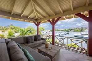 Ein überdachter Außenbereich mit einer Couch und einem Tisch, mit Blick auf einen Yachthafen und Boote.