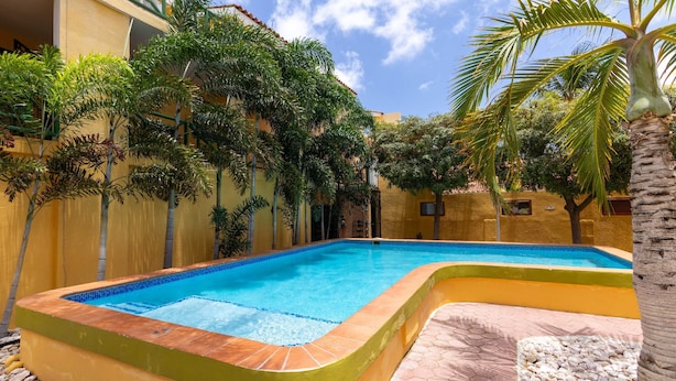 A swimming pool with a curved edge, surrounded by a yellow wall and palm trees. - Spanish Water Marina Quad Residences (Jan Thiel)