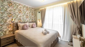 A bedroom with a floral wallpaper, a bed with a beige bedspread, a nightstand, a lamp, and a large window with curtains. - Lemair Luxury Apartments Kozani Deluxe Double Room (Kozani)