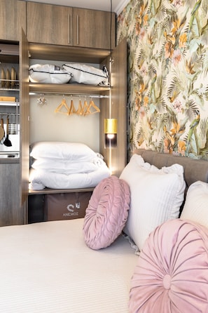 A bedroom with a wardrobe, a bed with pillows, a hanging rack, and a wall with a tropical wallpaper.
