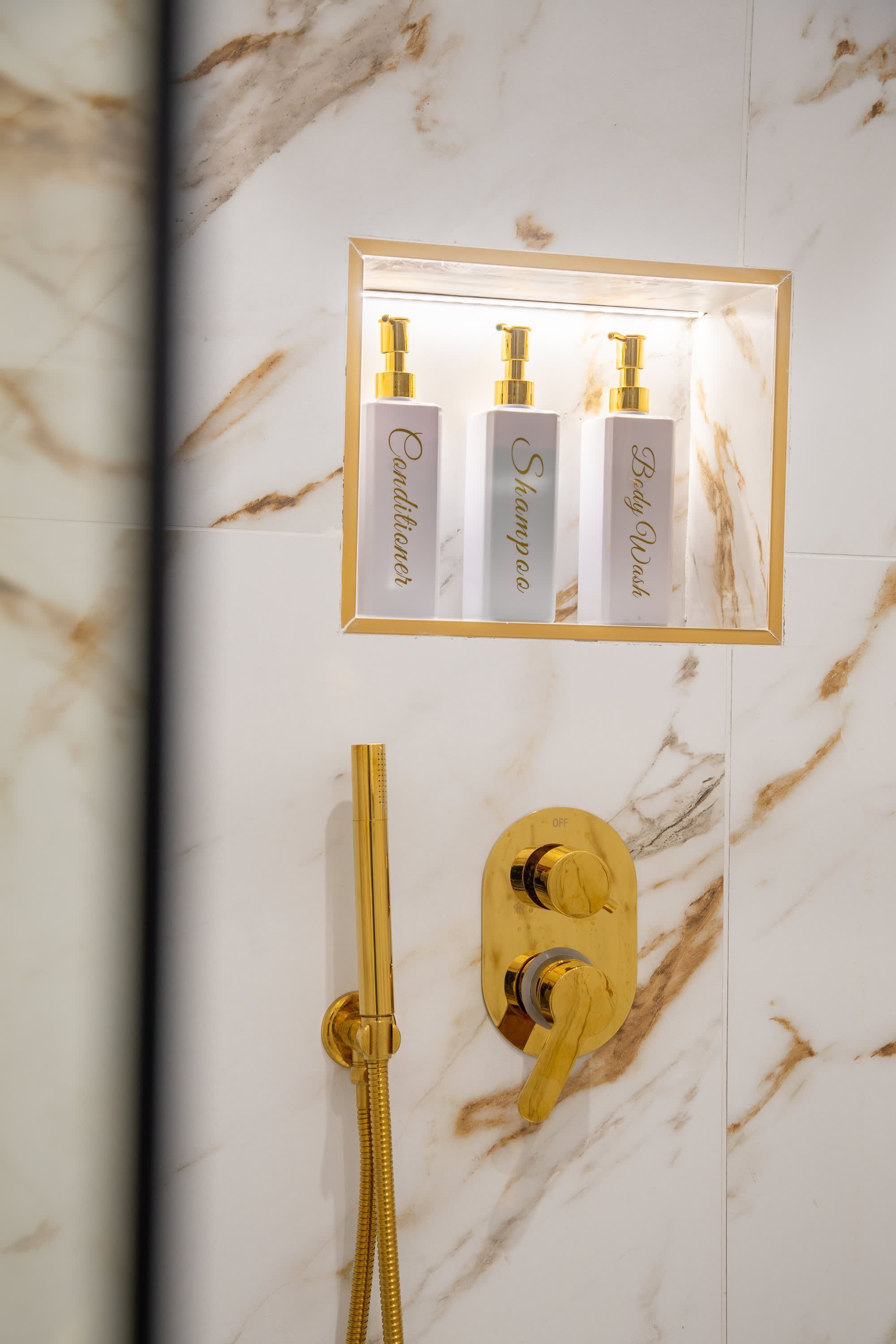 Une salle de bain avec un mur en marbre, une pomme de douche dorée et trois bouteilles de shampoing et d’après-shampoing.