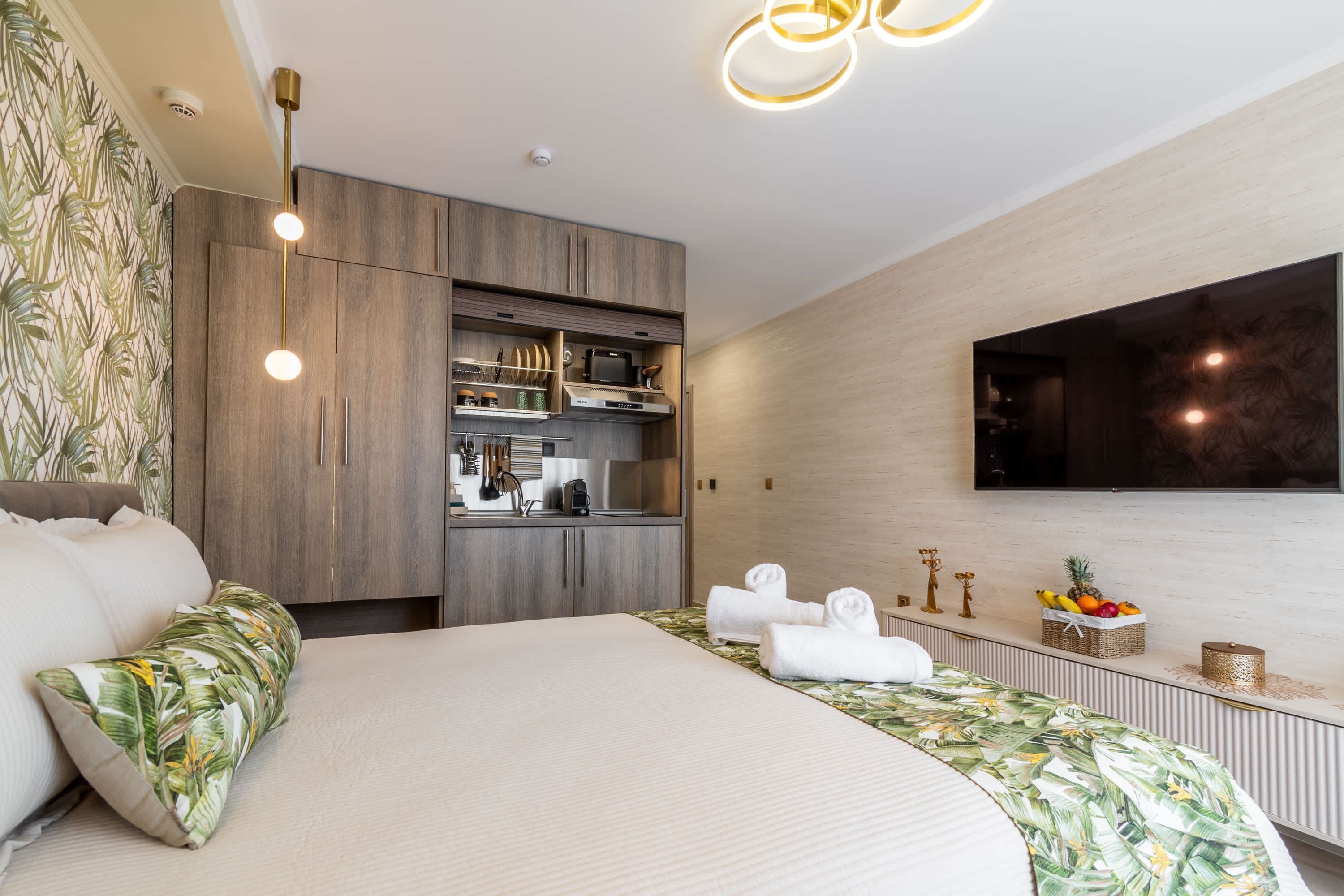 A modern hotel room with a large bed, a built-in kitchenette, a flat-screen TV, and a decorative wall with leaf patterns.