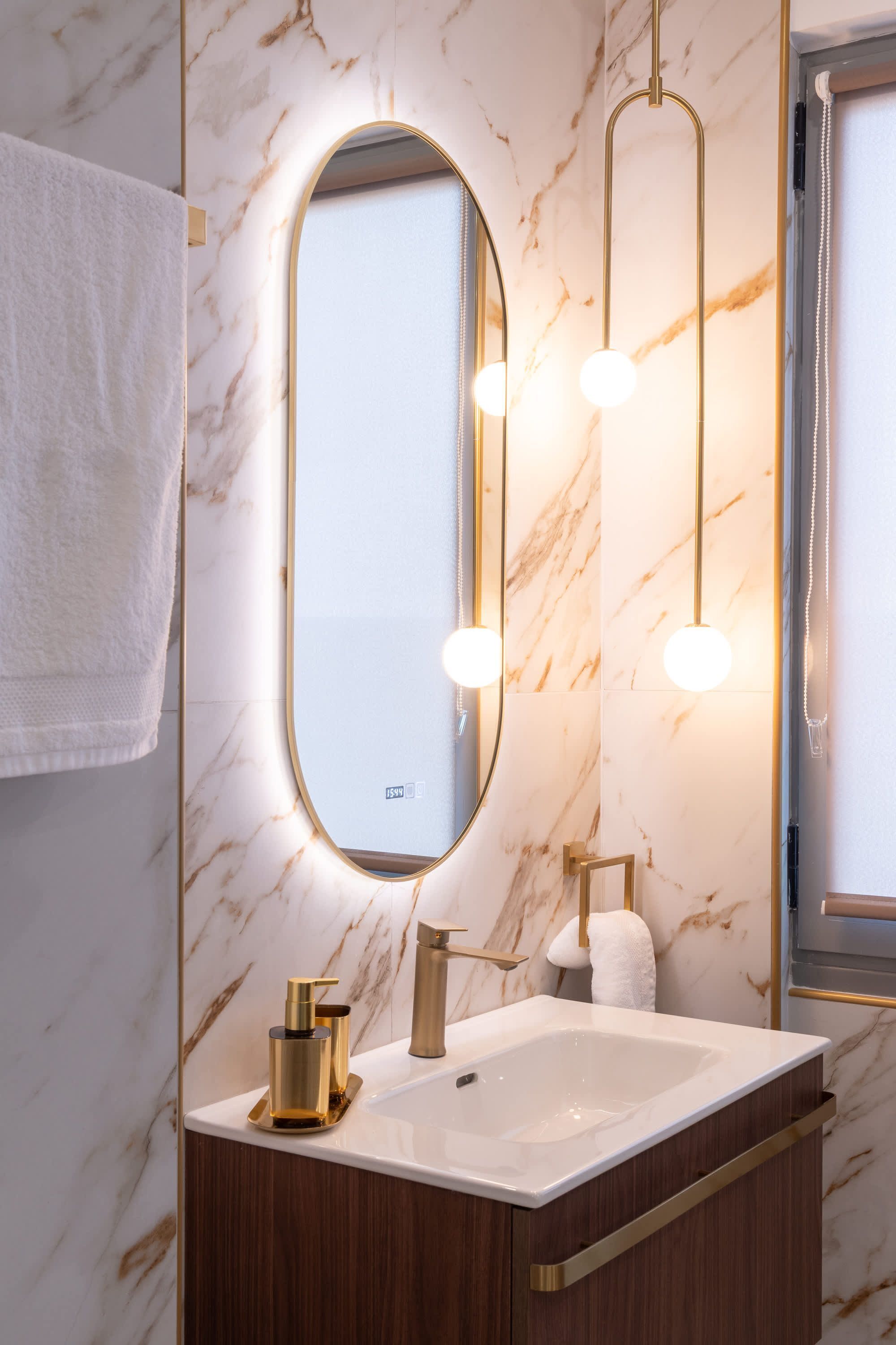 A bathroom with a marble wall, a large oval mirror, a white sink, gold fixtures, and a towel rack.